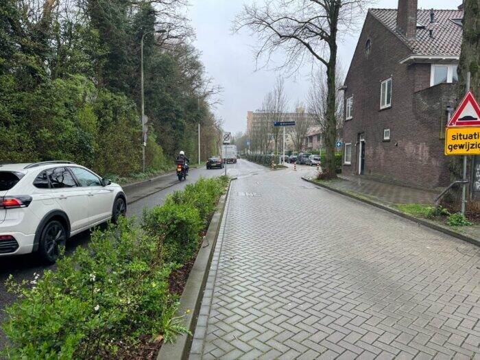 grijze autoweg met witte auto gescheiden van een grijs fietspad door middel van een groene struiken strook